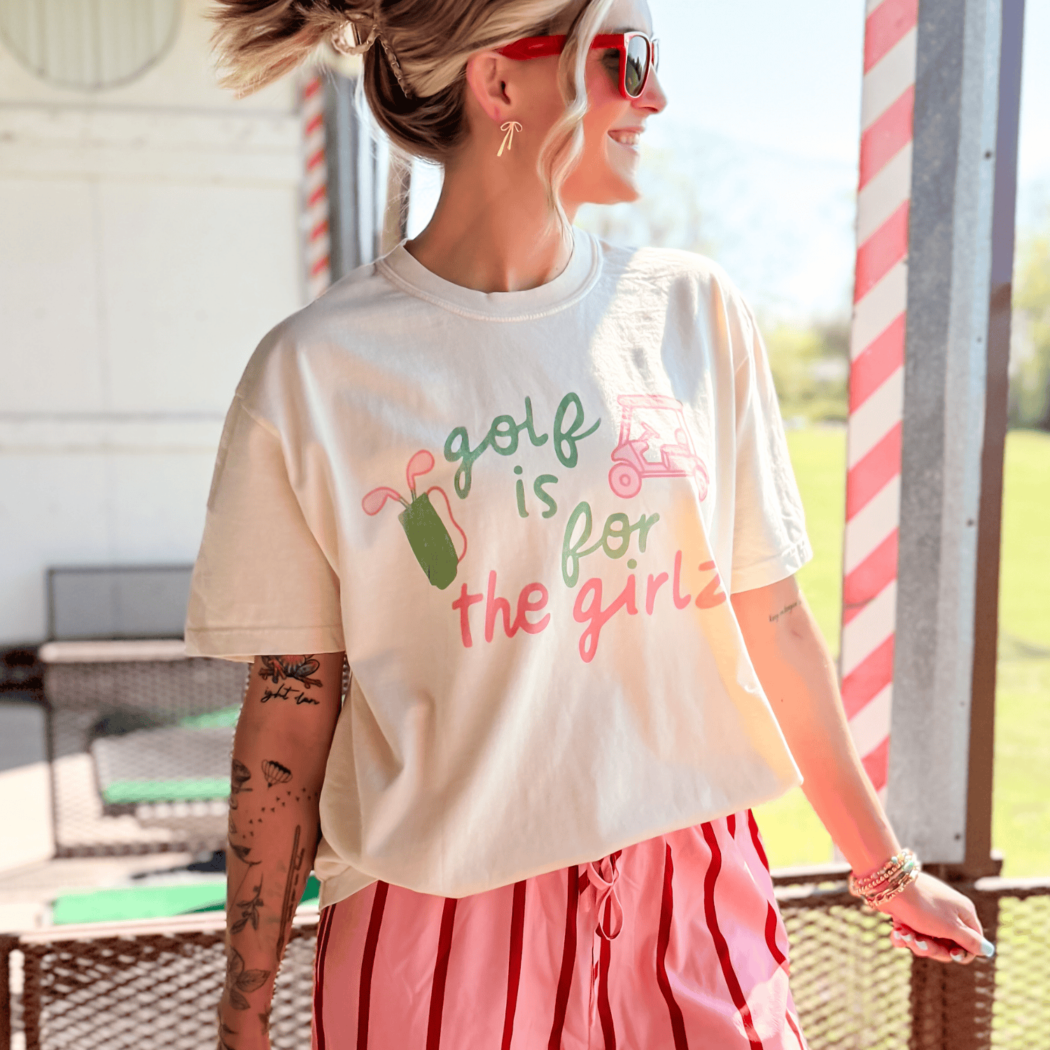 Woman wearing a t-shirt with 'Golf is for the girls' text and graphics outdoors.