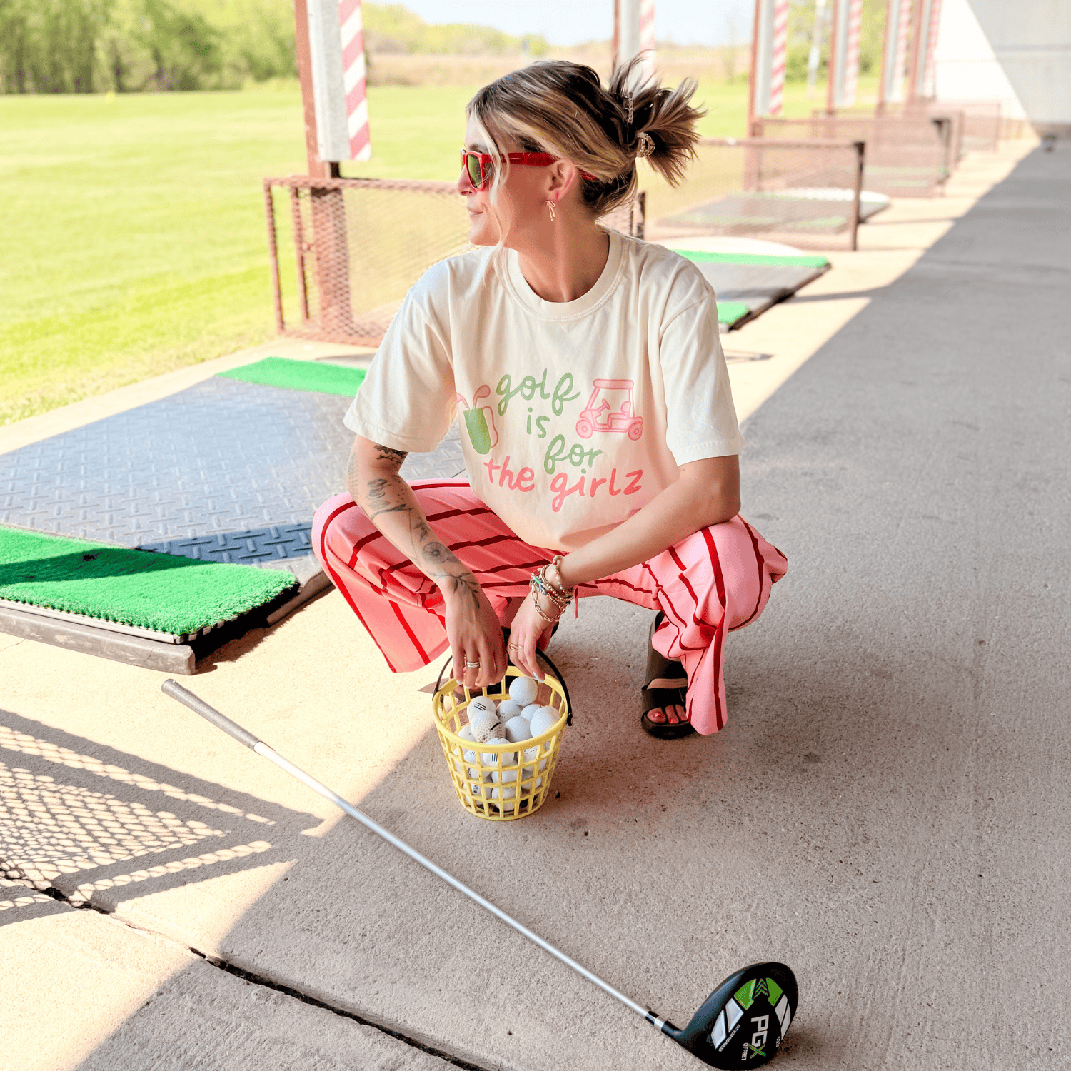 Person at a driving range with a golf club and basket, wearing a t-shirt with text.
