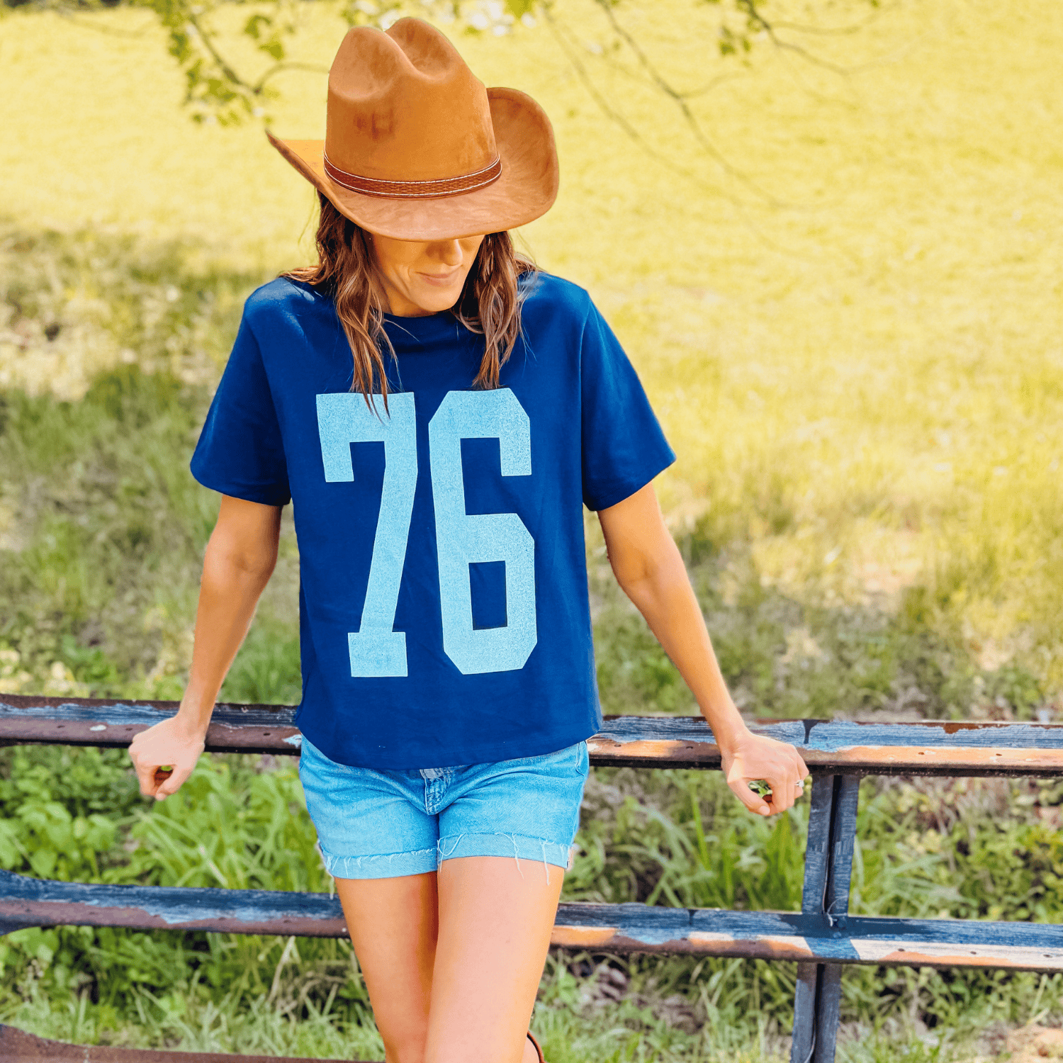 Navy distressed 76 mid tee worn by a woman with a brown hat, standing by a fence in a grassy field.