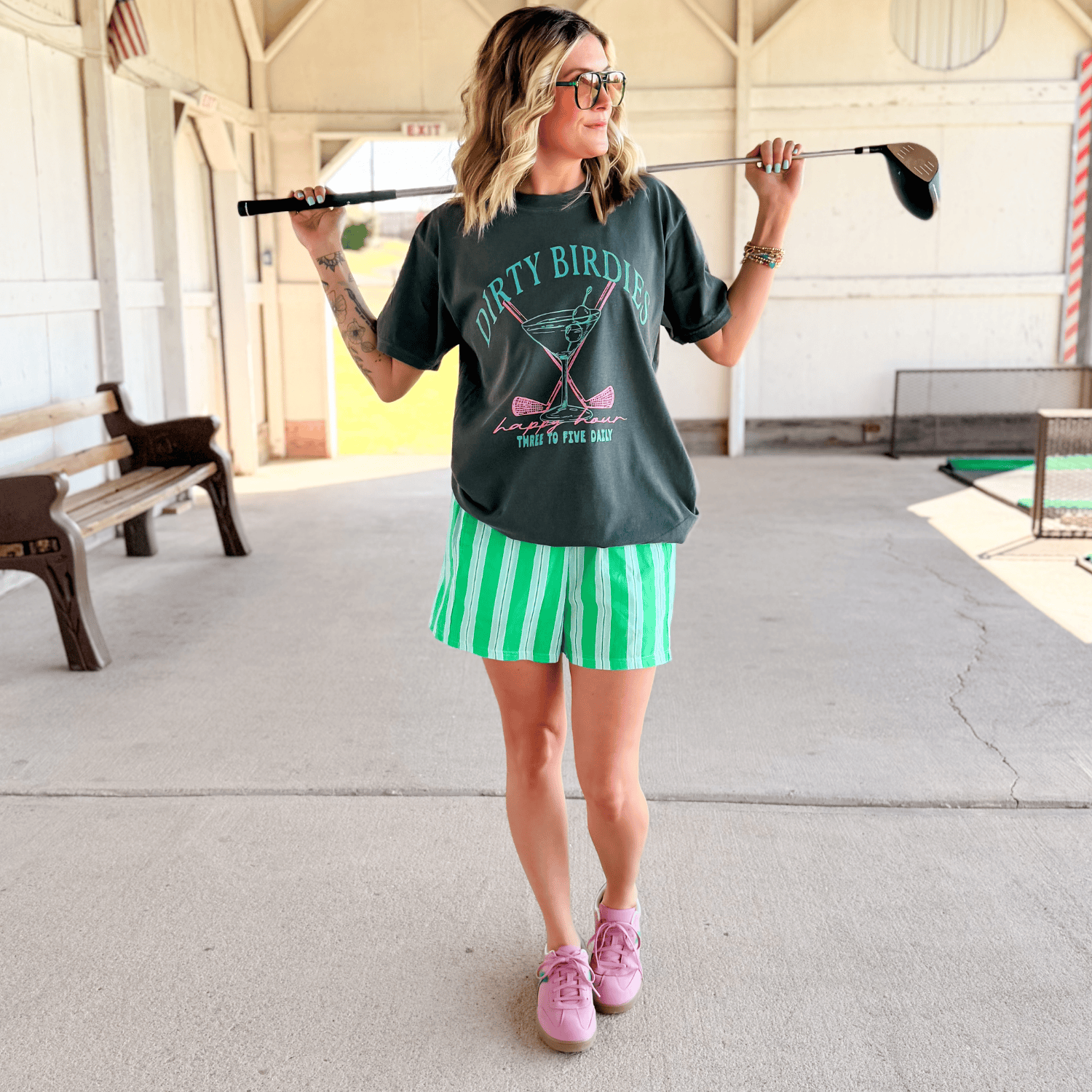 Woman holding a golf club wearing a casual outfit with a graphic t-shirt and striped shorts.