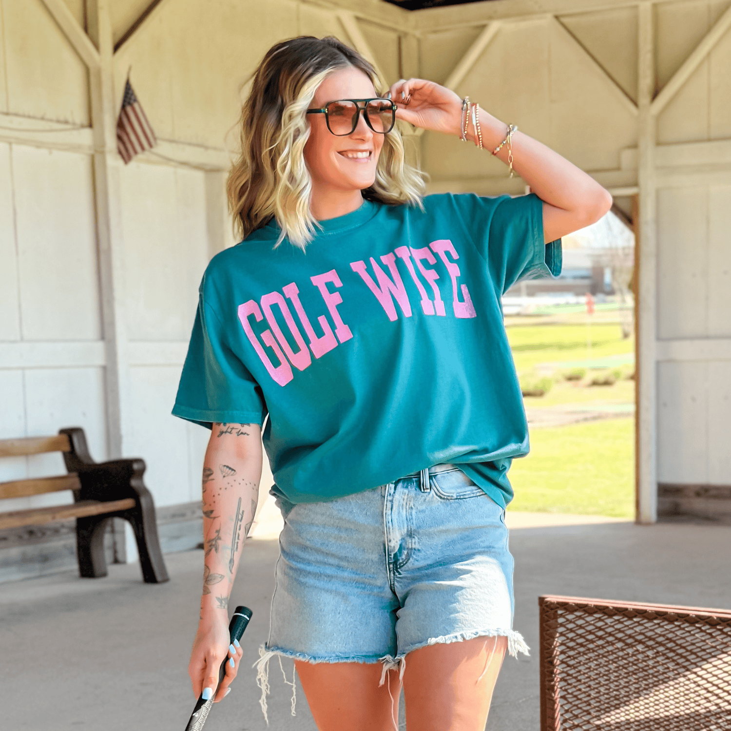 Woman wearing a teal 'GOLF WIFE' shirt outdoors.
