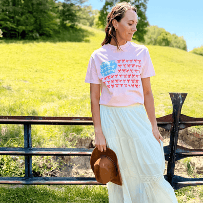 American Flag Bows Tee in pink with blue and red bow patterns, worn by a woman standing near a wooden fence.