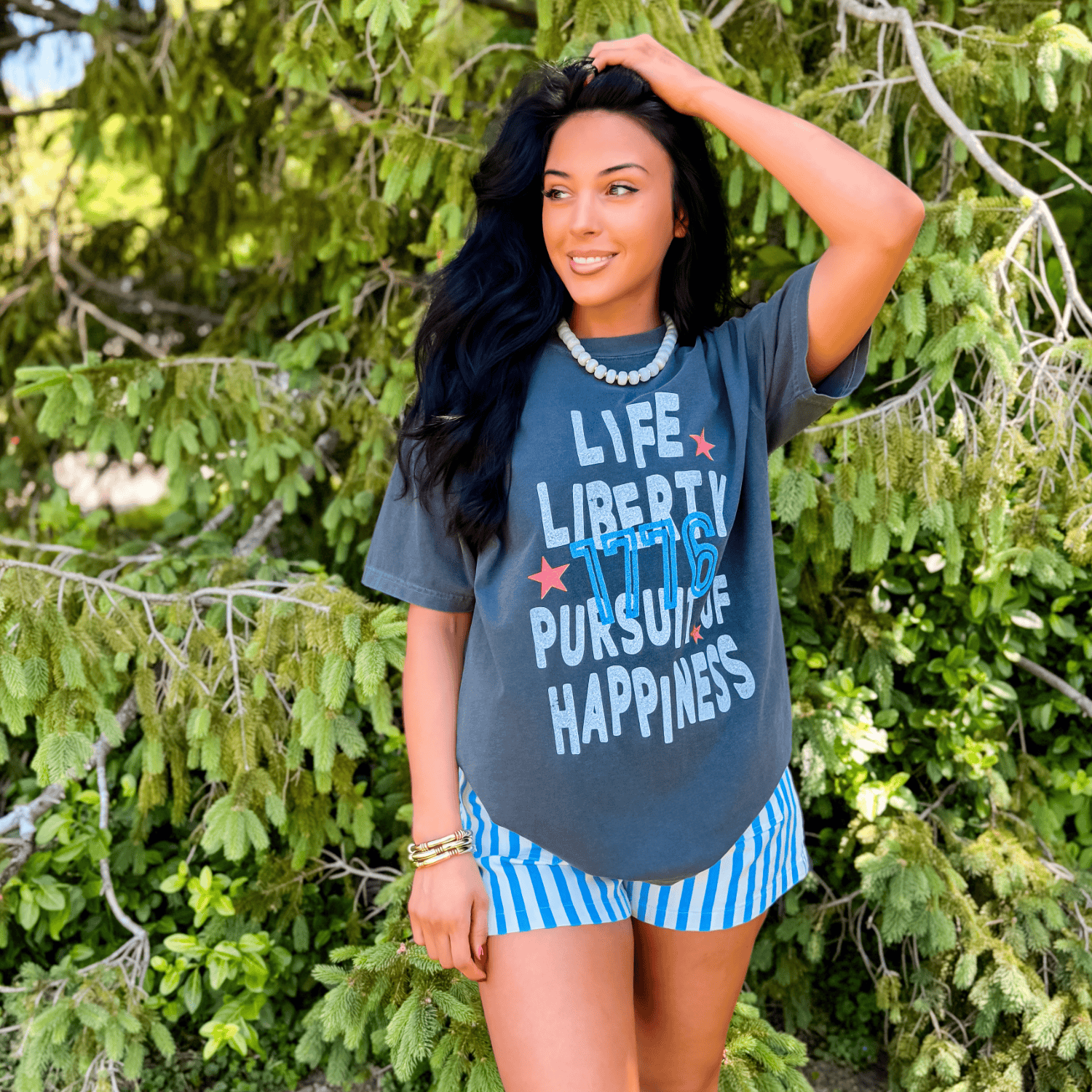 Pursuit of Happiness tee in charcoal gray with colorful text, worn by a woman standing among green foliage.