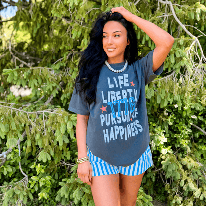 Pursuit of Happiness tee in charcoal gray with colorful text, worn by a woman standing among green foliage.