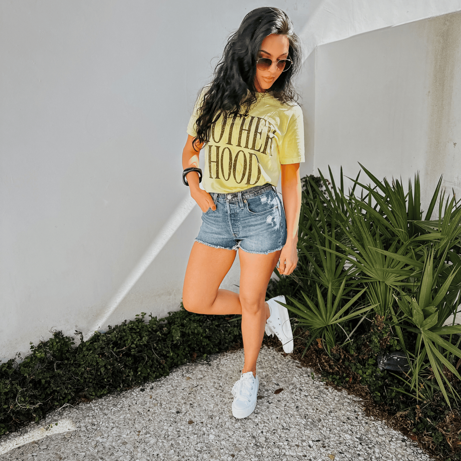 Bold Motherhood banana tee in yellow, paired with denim shorts and white sneakers, standing against a white wall.