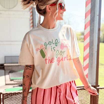 Woman wearing a t-shirt with 'Golf is for the girls' text and graphics outdoors.