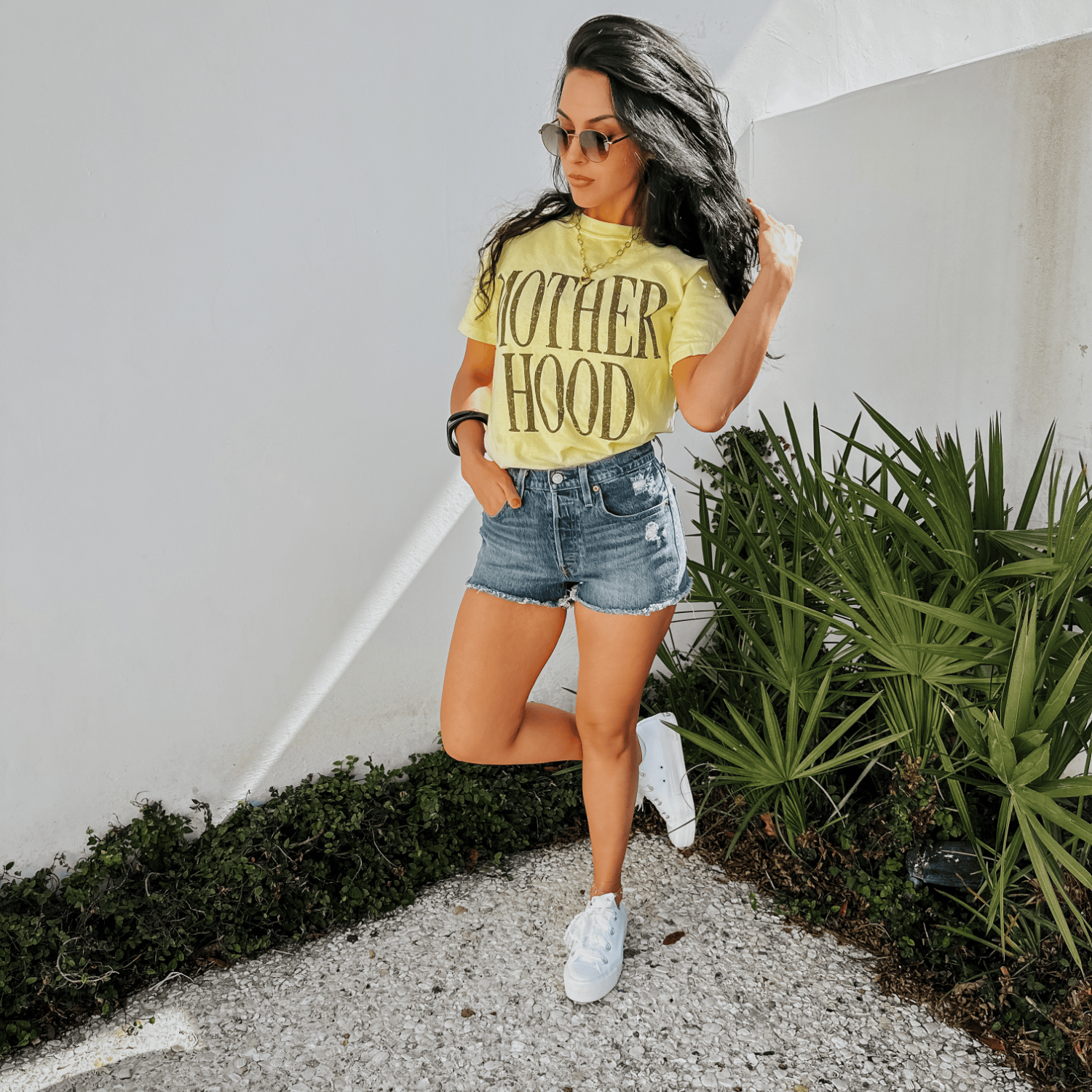 Bold Motherhood banana tee in yellow, paired with denim shorts and white sneakers, model poses against a white wall.