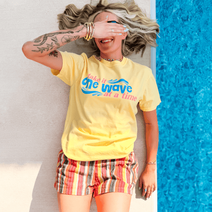 Yellow One Wave Tee with a colorful graphic, worn by a smiling person lying beside a blue pool.