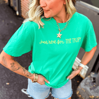 Green t-shirt with "just here for THE TROPES" in yellow text, styled with denim shorts and accessories.