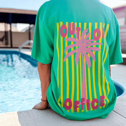 Neon Out of Office tee in green with a pink palm tree graphic and yellow stripes, worn by a person near a pool.