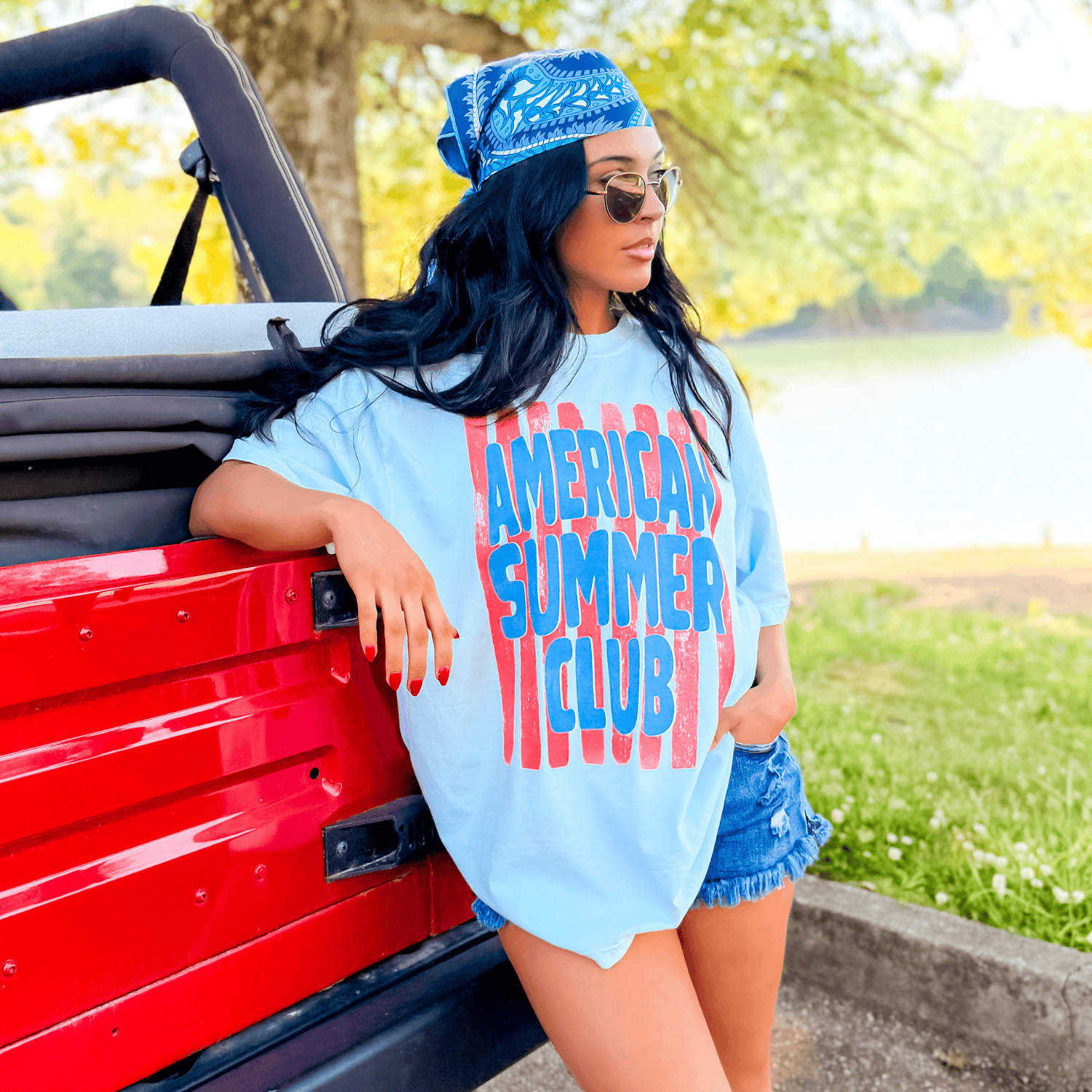 Light blue American Summer Club tee with bold red lettering, worn by a woman beside a red vehicle in a sunny outdoor setting.